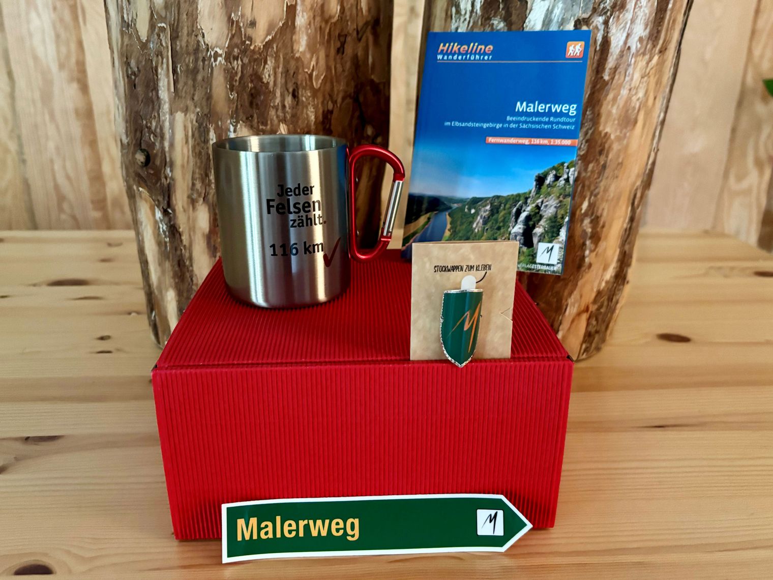 Geschenkbox Malerweg Auf einem Holztisch steht eine rote Box, darauf steht eine Silberne Tasse, ein Stocknagel, ein Buch und davor ein kleines grünes Schild mit dem Text "Malerweg".
