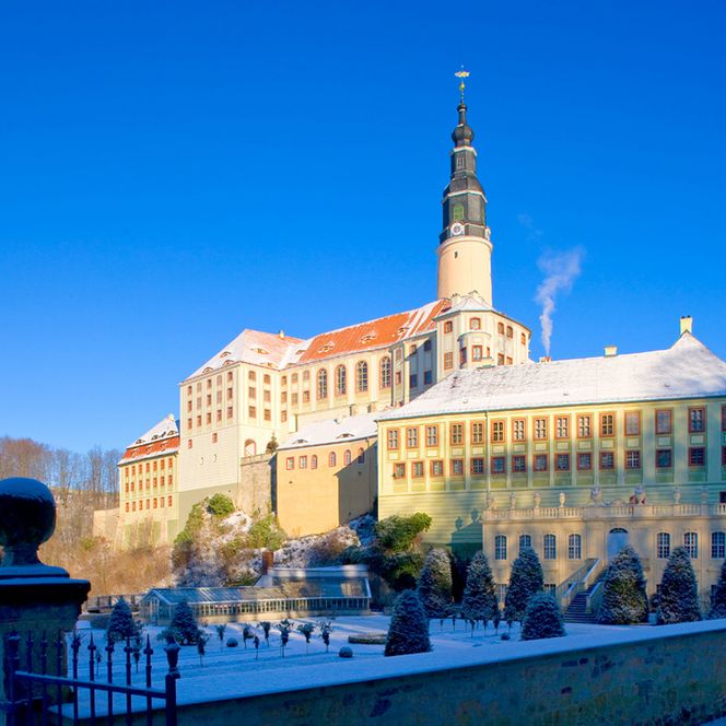 Schneebedecktes Schloss auf einem Hügel, umgeben von Bäumen und klarem blauen Himmel.