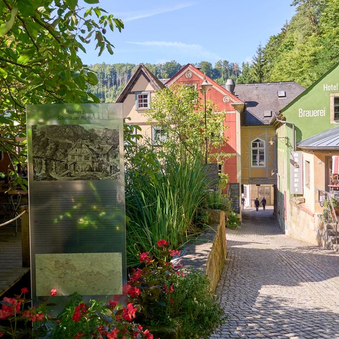 Bunte Häuser und ein Weg, umgeben von Bäumen und Blumen, unter blauem Himmel.