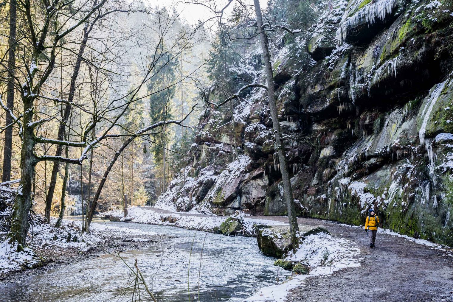 Wanderer auf einem schneebedeckten Weg entlang eines gefrorenen Flusses, umgeben von Felsen und Bäumen.