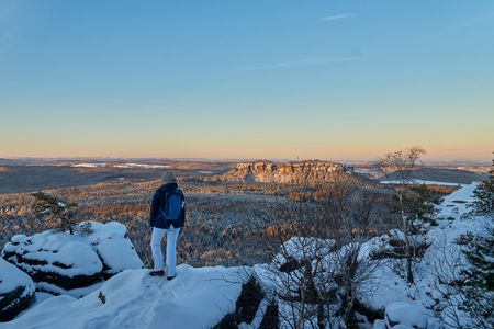 Person in blauer Jacke steht auf schneebedecktem Felsen und blickt auf eine winterliche Landschaft.