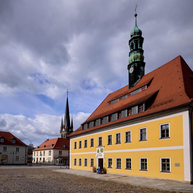 Gelbes Gebäude mit rotem Dach und Uhrturm, umgeben von grauem Himmel und Wolken.