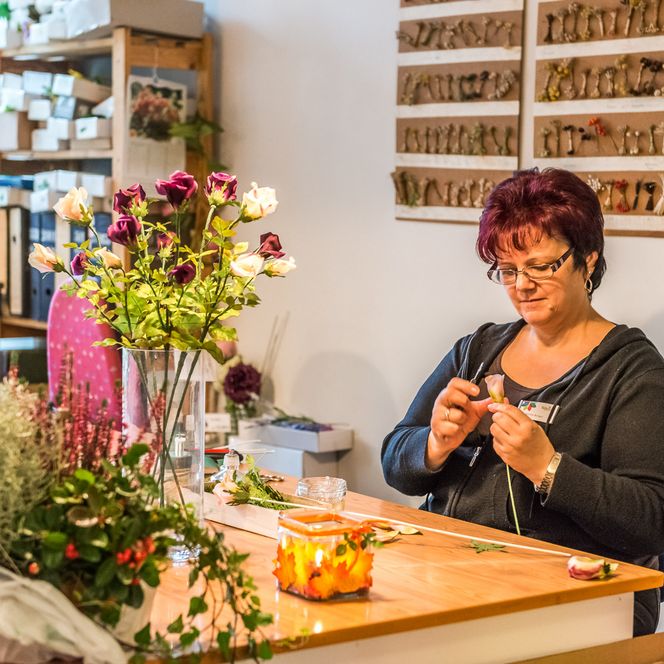 Frau mit Brille sitzt an einem Tisch und arrangiert Blumen, umgeben von Pflanzen und Werkzeugen.