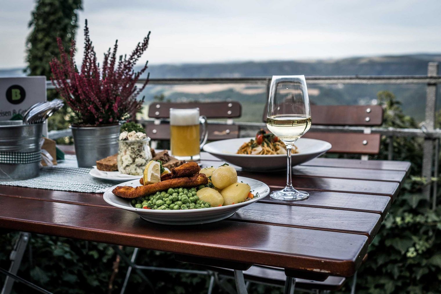 Zwei Teller mit Essen, ein Glas Bier und ein Glas Wein auf einem Holztisch mit Aussicht.