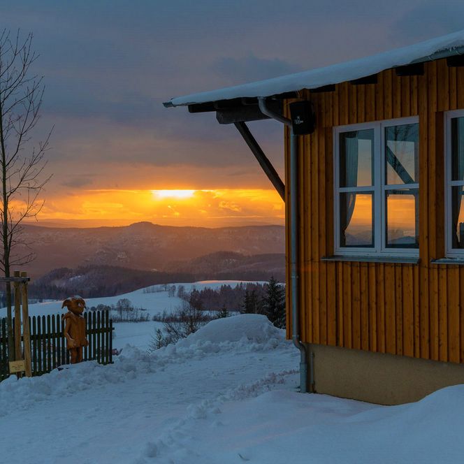 Holzhütte im Schnee mit Blick auf eine winterliche Landschaft und Sonnenuntergang im Hintergrund.