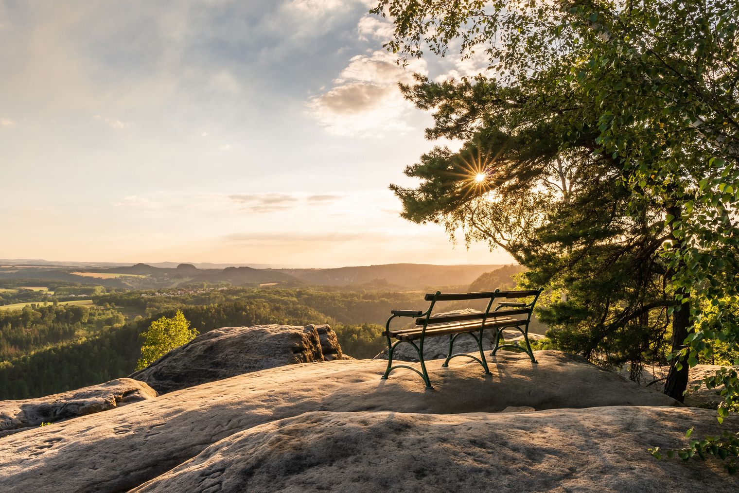 Blick auf eine Landschaft mit einer Bank auf einem Felsen, Sonnenuntergang im Hintergrund.