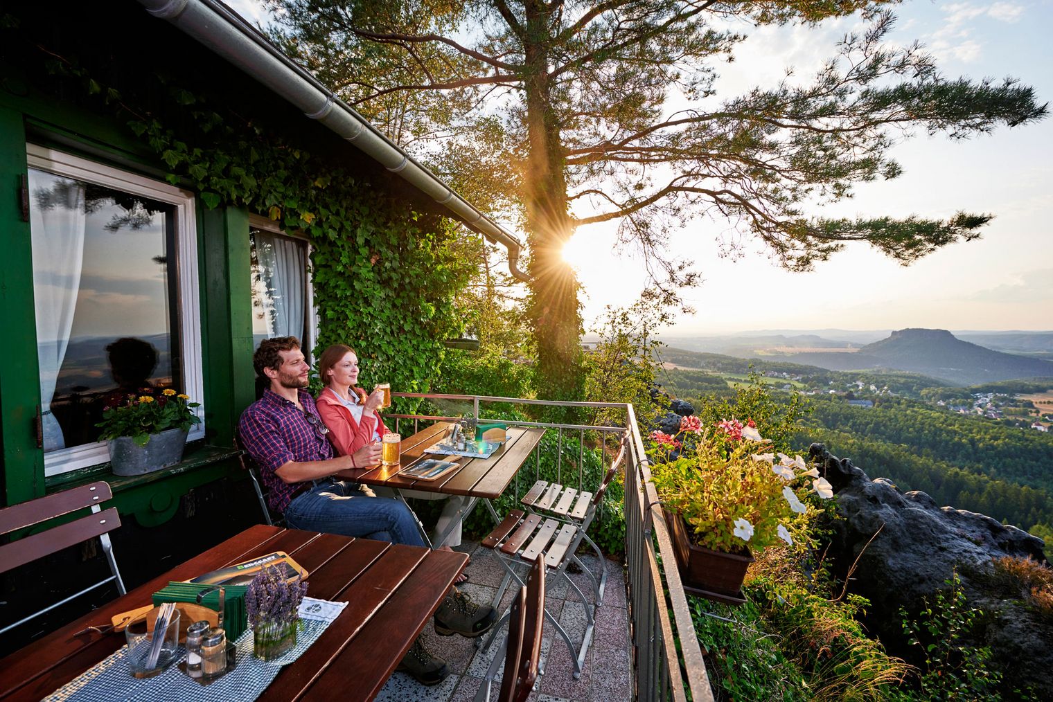 Paar sitzt auf einer Terrasse der Wirtschaft auf dem Papststein mit Blick auf die Landschaft bei Sonnenuntergang, Getränke und Snacks auf dem Tisch.