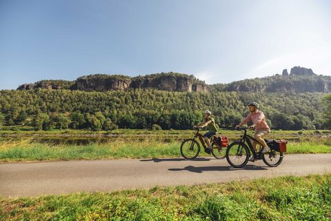Zwei Personen fahren auf Fahrrädern entlang der Elbe in einer grünen Landschaft mit Hügeln im Hintergrund.