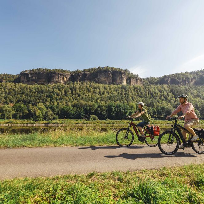 Zwei Personen fahren auf Fahrrädern entlang der Elbe in einer grünen Landschaft mit Hügeln im Hintergrund.