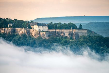 Burg auf einem Hügel, umgeben von Nebel und sanften Hügeln im Hintergrund.