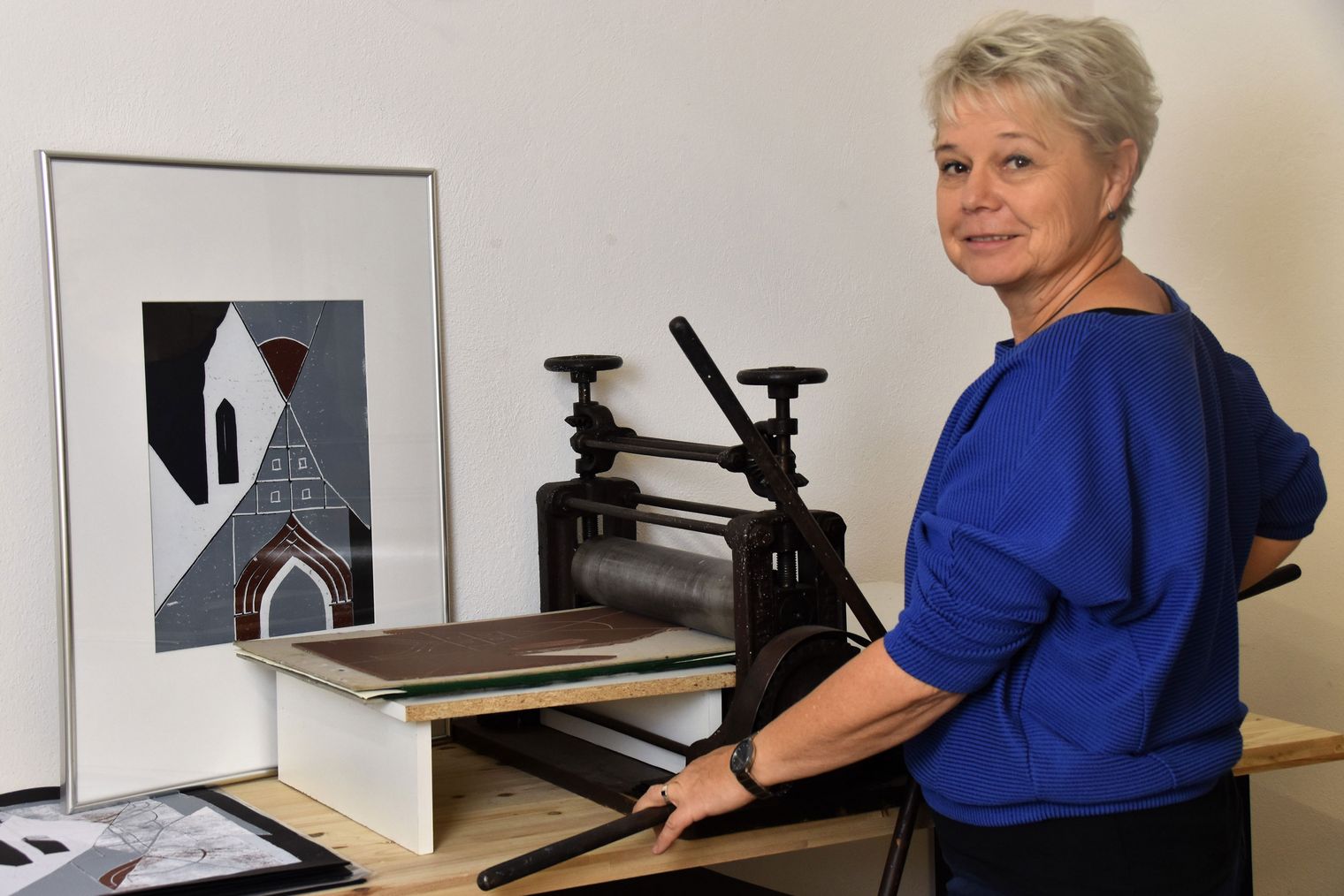 Künstlerin mit kurzem, grauem Haar steht neben einer Druckmaschine, umgeben von Druckmaterialien und einem gerahmten Kunstwerk.