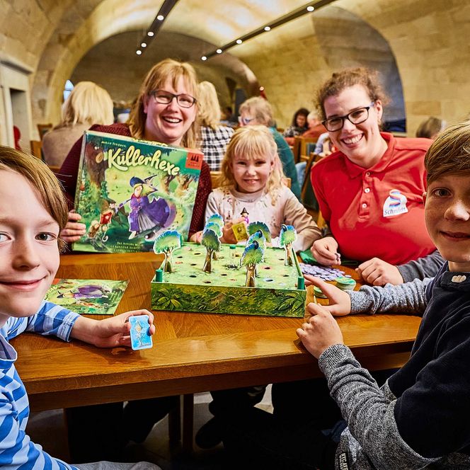 Fünf Kinder und Erwachsene sitzen an einem Tisch mit einem Brettspiel und lächeln in die Kamera.