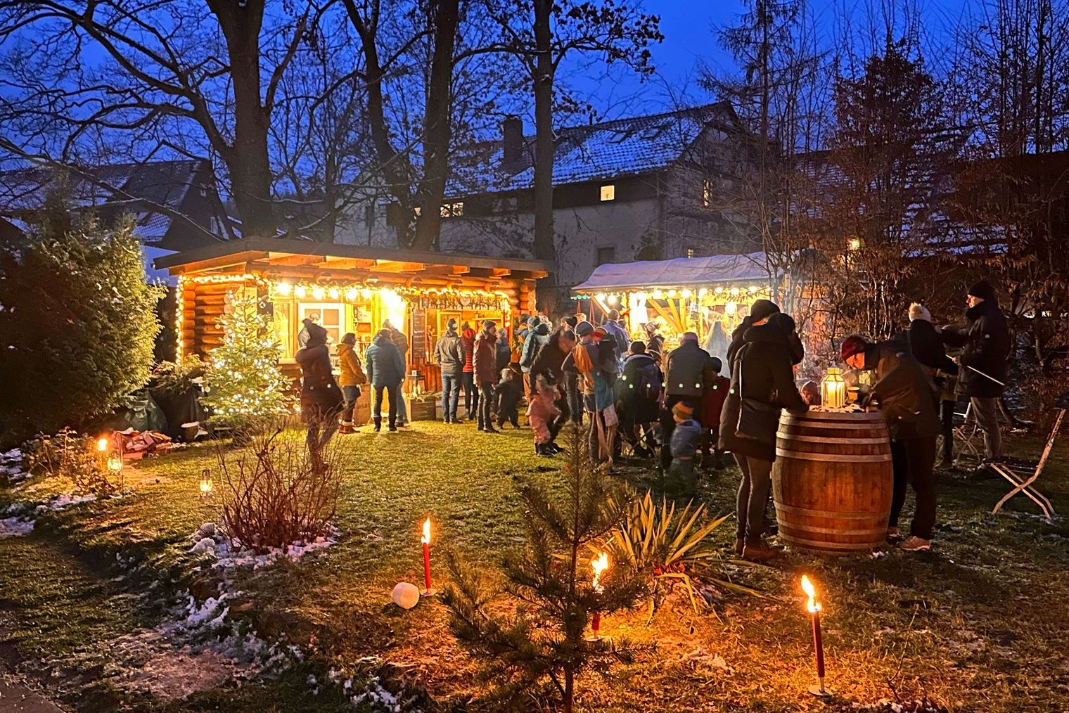 Beleuchteter Weihnachtsmarkt mit Menschen, Tannenbäumen und festlicher Dekoration bei Dämmerung.