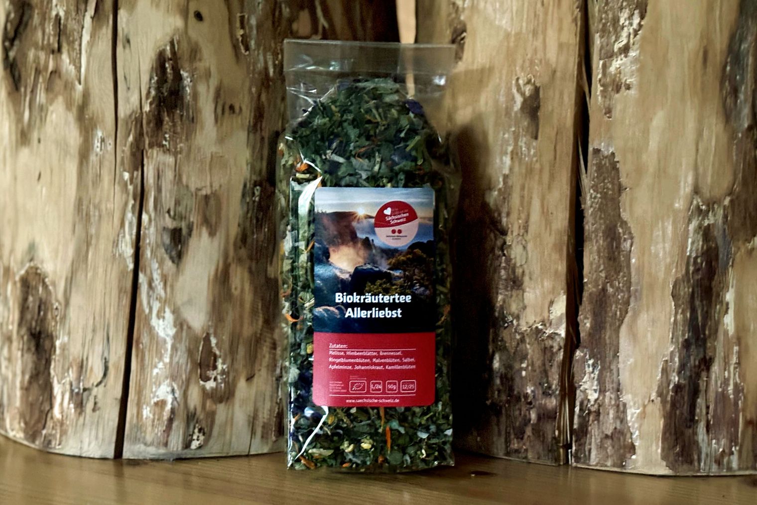 Packung Biokräutertee "Allerliebst" Vor einem Holzstamm steht eine durchsichtige Packung mit Teekräutern, darauf ein Foto von Felsen bei Sonnenaufgang.