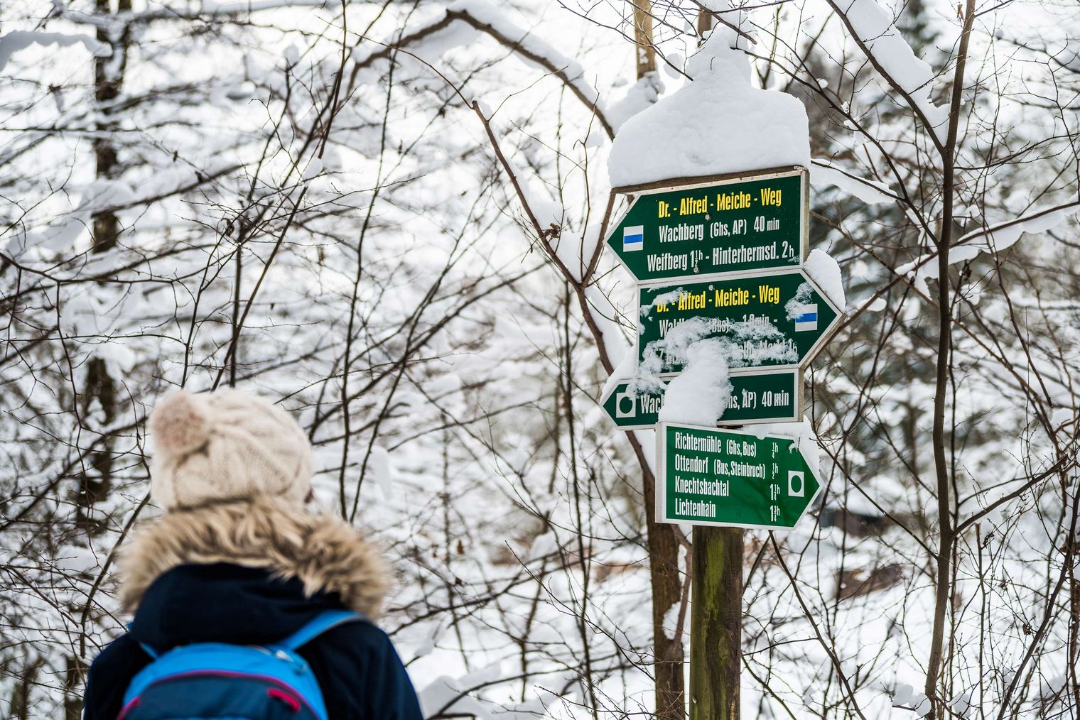 Wanderer mit Mütze steht vor einem beschneiten Wegweiser im Wald.