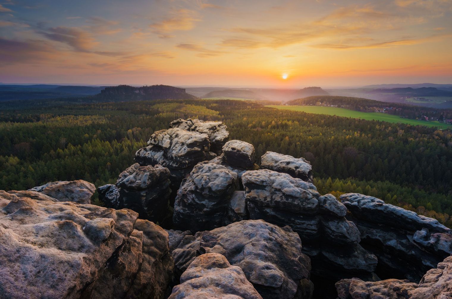Sonnenaufgang über einer bewaldeten Landschaft mit Felsen im Vordergrund.
