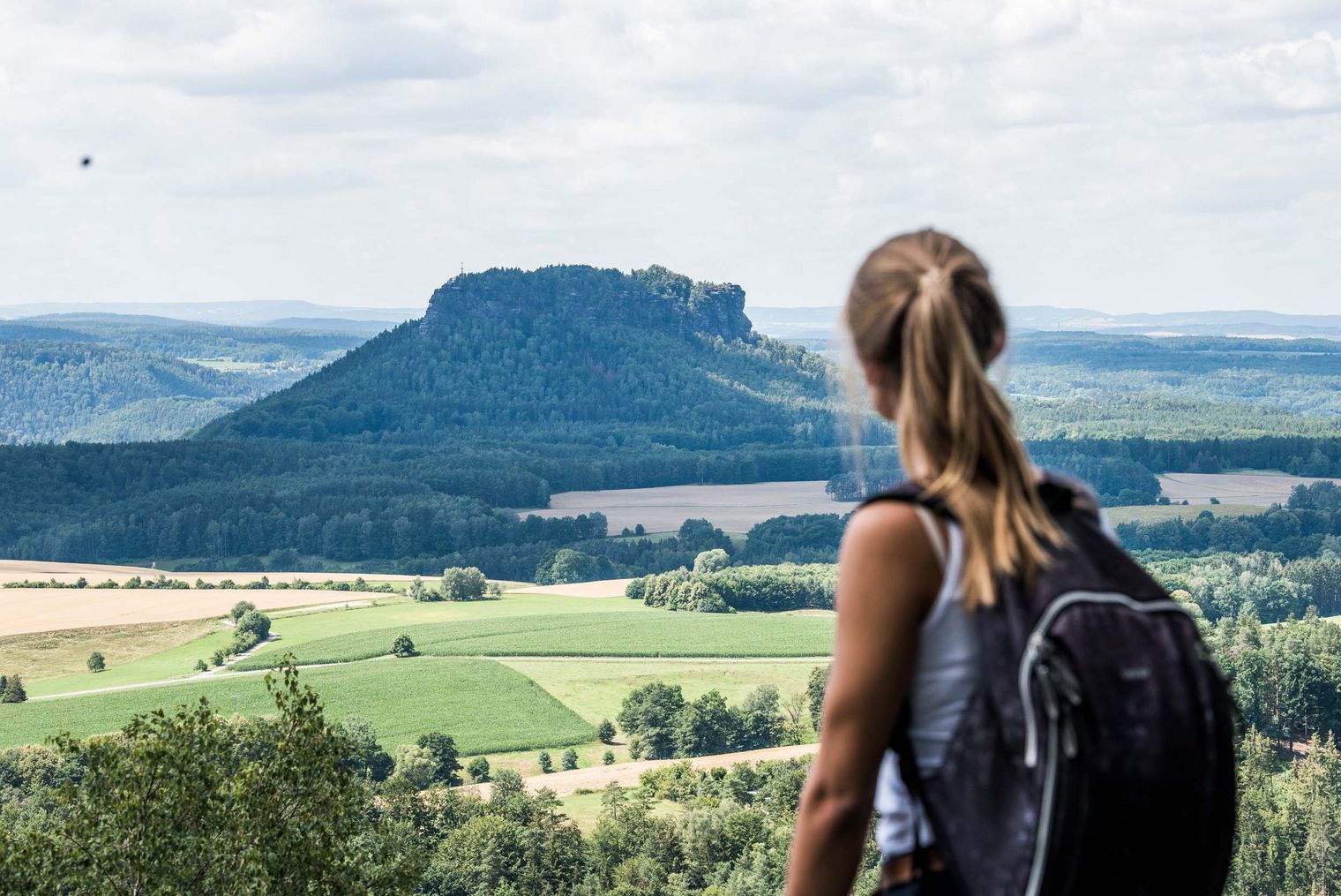 Frau mit Rucksack blickt auf eine hügelige Landschaft mit einem markanten Felsen, dem Lilienstein, im Hintergrund.