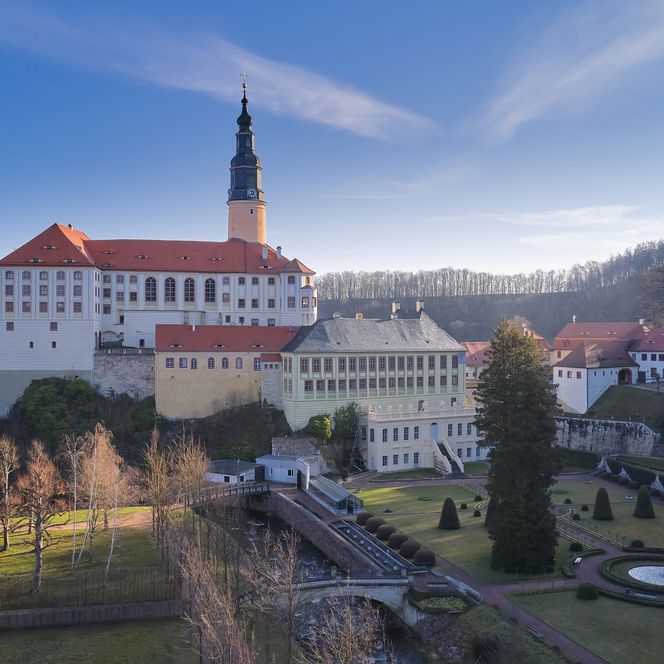 Historisches Gebäude (Schloss Weesenstein) mit Turm und roten Dächern, umgeben von Bäumen und einem klaren Himmel.