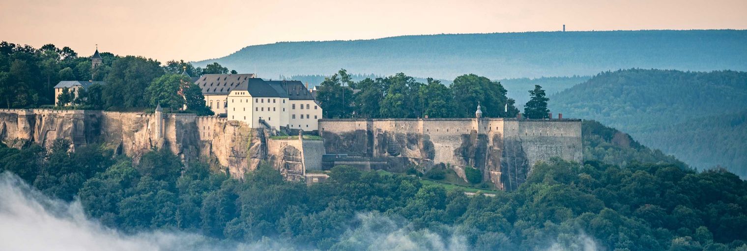 Blick auf eine historische Festung Königstein auf einem Felsplateau, umgeben von Bäumen und Nebel