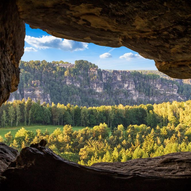 Blick aus einer Höhle auf eine grüne Landschaft mit Bergen im Hintergrund und blauem Himmel.