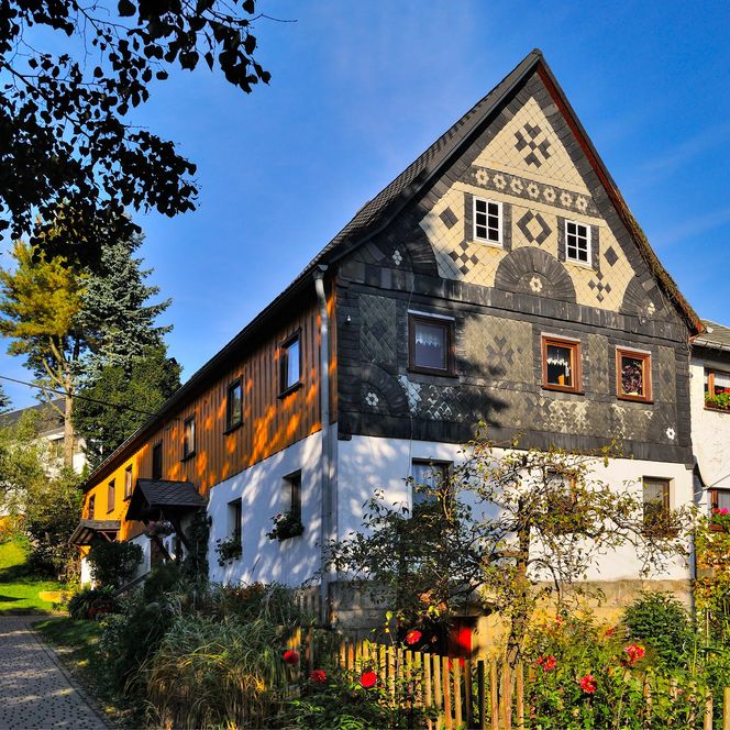Fachwerkhaus mit bunten Fensterläden und Garten, umgeben von Bäumen und blauem Himmel.