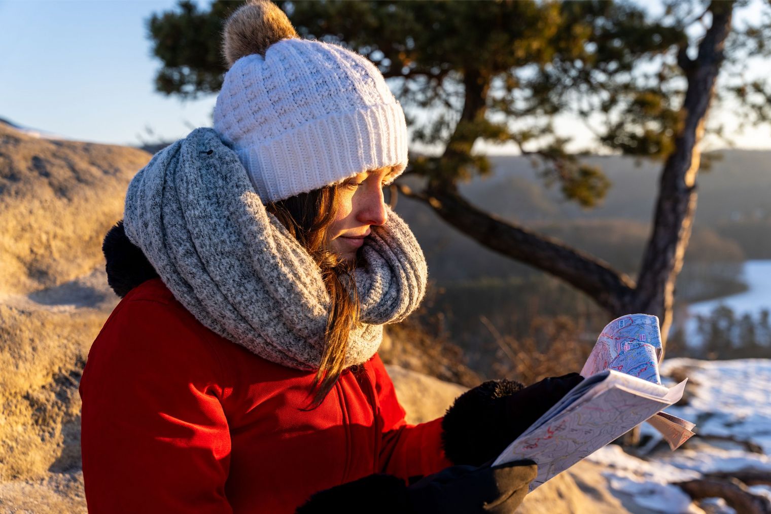 Frau in rotem Mantel und weißer Mütze liest ein Buch im Freien, umgeben von Natur.