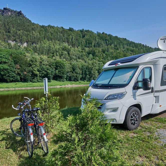 Wohnmobil steht am Ufer eines ruhigen Gewässers, im Hintergrund eine bewaldete Hügelkette. Zwei Fahrräder lehnen an e...