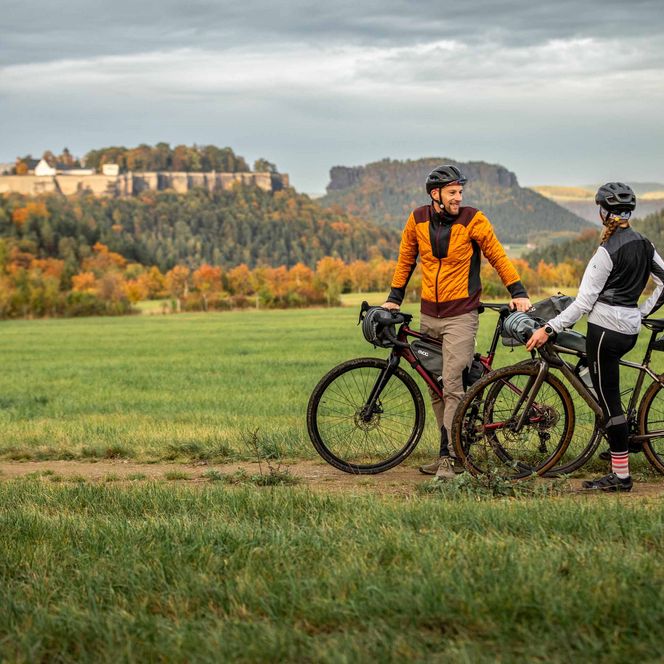 Zwei Radfahrer stehen auf einem Feldweg, umgeben von grünen Wiesen und Bergen im Hintergrund.