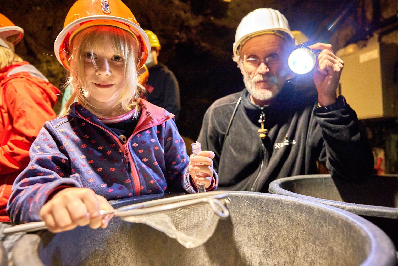 Ein Mädchen mit einem orangefarbenen Helm hält ein Netz, während ein Mann mit einer Taschenlampe daneben steht.