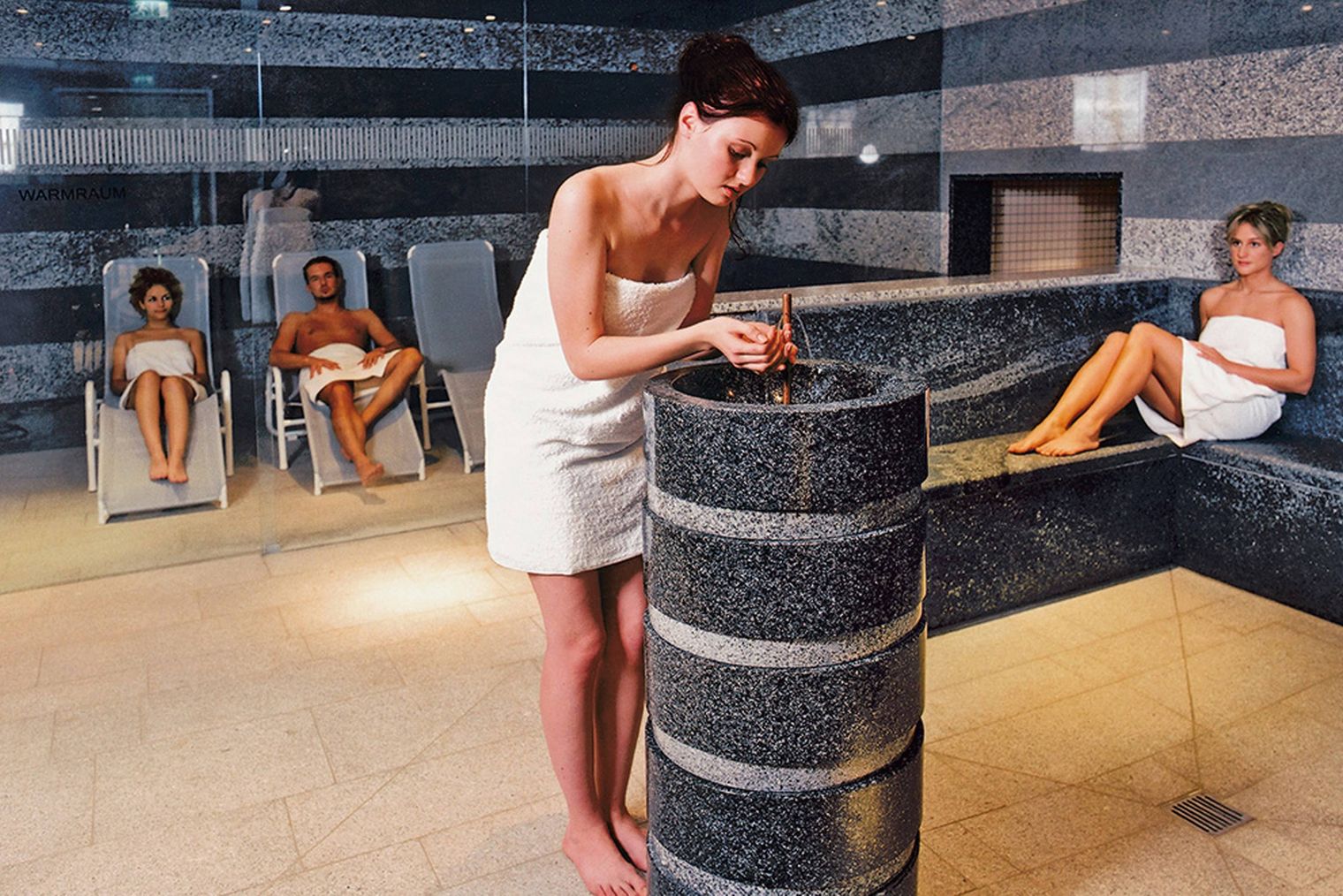 Drei Frauen in Handtüchern in einem Spa-Bereich, eine Frau beugt sich über einen Steintisch.