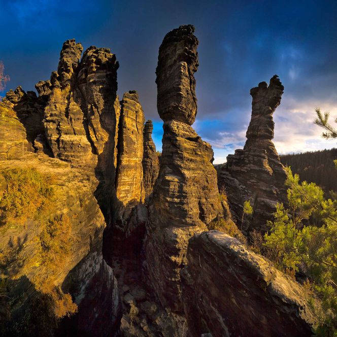 Herkulessäulen im Bielatal: Felsformationen mit spitzen Gipfeln, umgeben von grüner Vegetation und dramatischem Himmel.