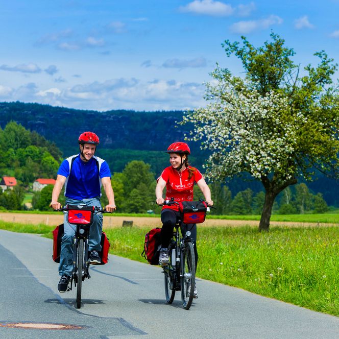 Zwei Radfahrer in blauen und roten Shirts fahren auf einem Weg, umgeben von grünen Wiesen und Bäumen.