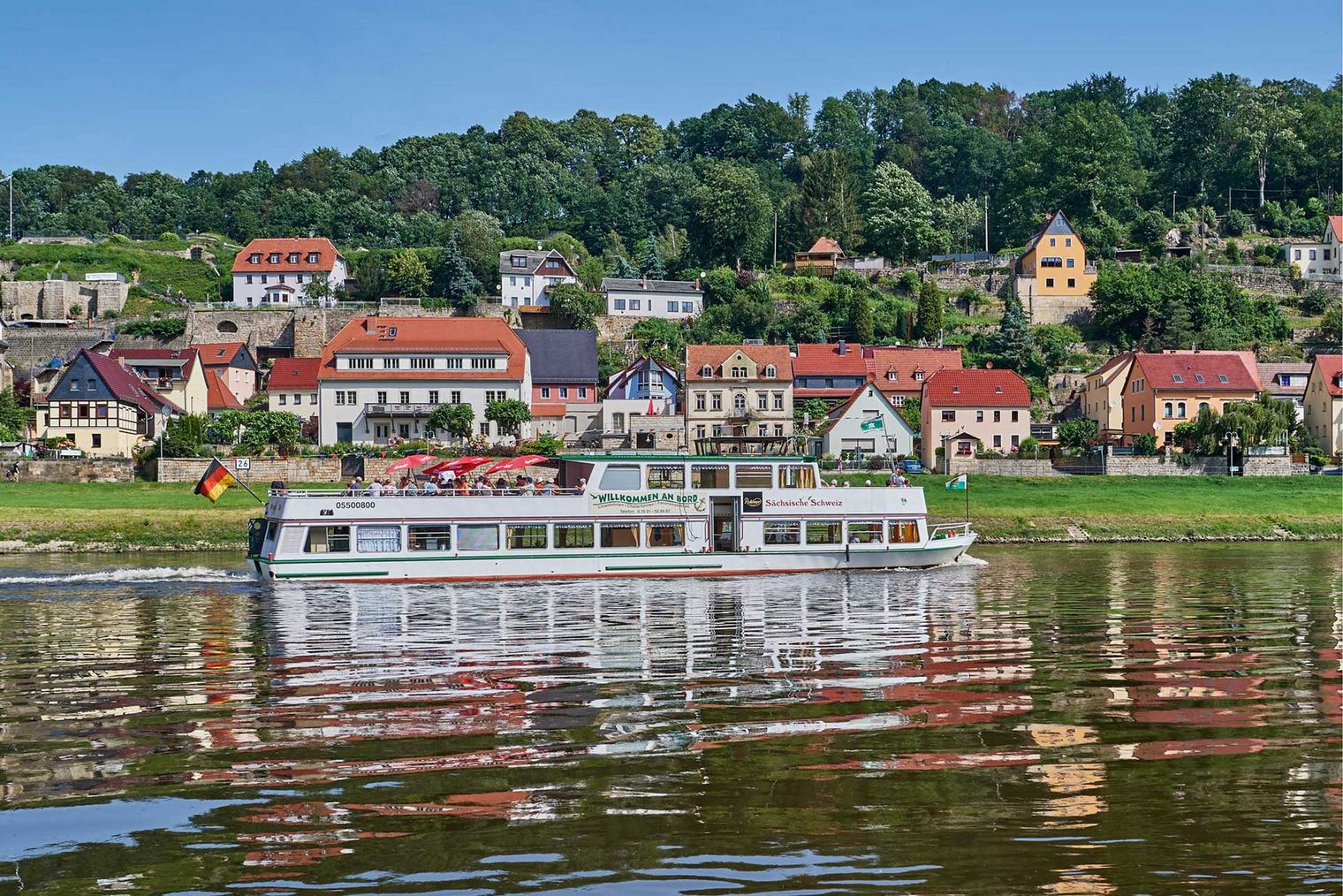 Ein Schiff fährt auf einem ruhigen Fluss, im Hintergrund sind Häuser mit roten Dächern und grüne Hügel.