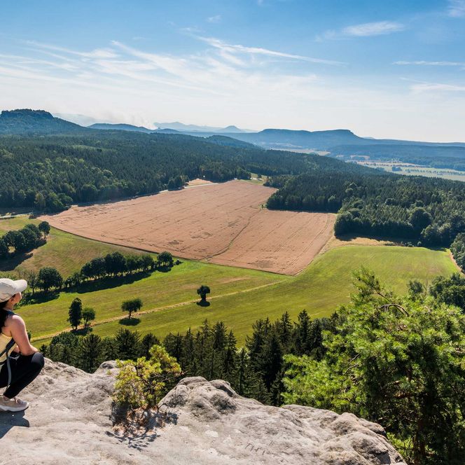 Wanderer sitzt auf einem Felsen und blickt auf eine grüne Landschaft mit Feldern und Hügeln.