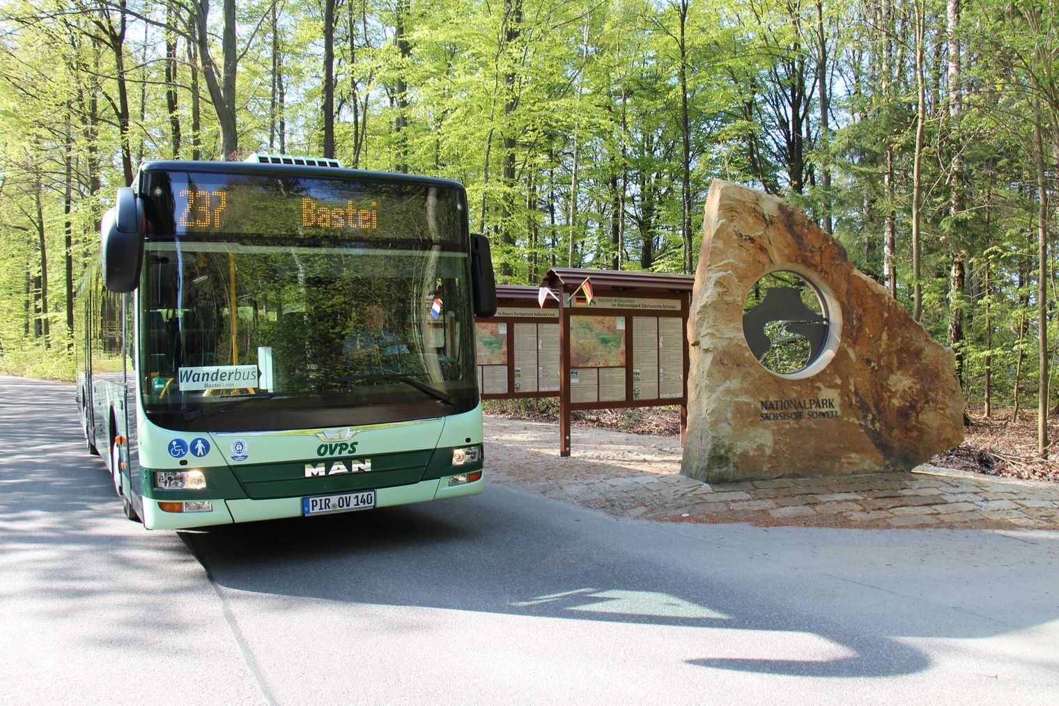 Grüner WanderBus fährt an einer Haltestelle mit Wartehäuschen und Bäumen vorbei.