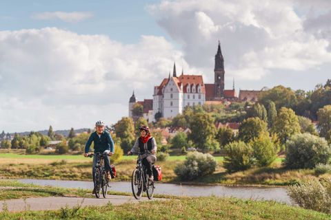 Zwei Radfahrer fahren am Fluss entlang, im Hintergrund Meißen mit der Albrechtsburg Burg und bewaldete Hügel.