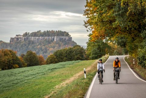 Zwei Radfahrer mit Gravelbike auf einer Straße, umgeben von Bäumen und Wiesen, im Hintergrund die Festung Königstein.