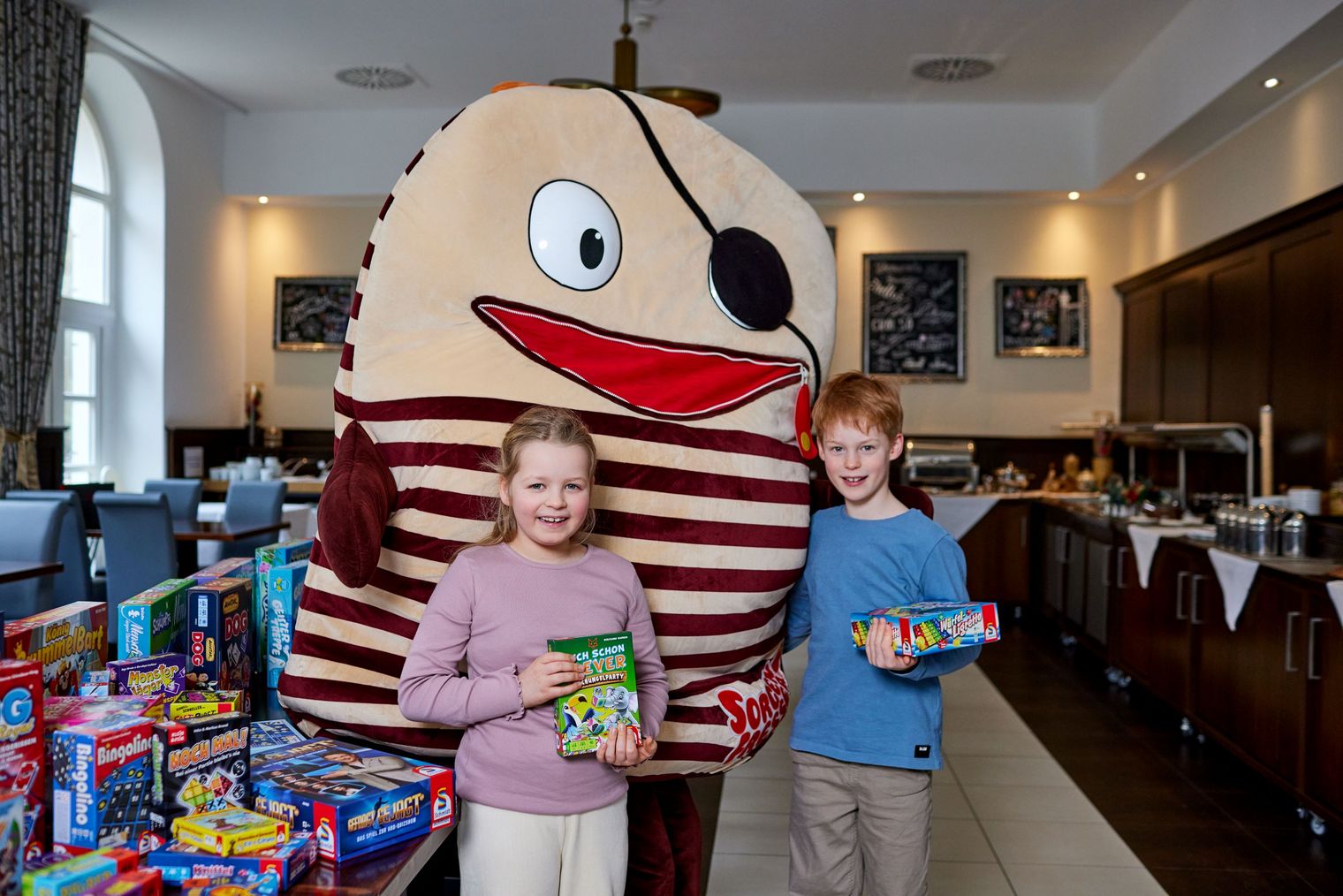 Zwei Kinder stehen neben einem großen, gestreiften Maskottchen mit Augenklappe und breitem Lächeln.