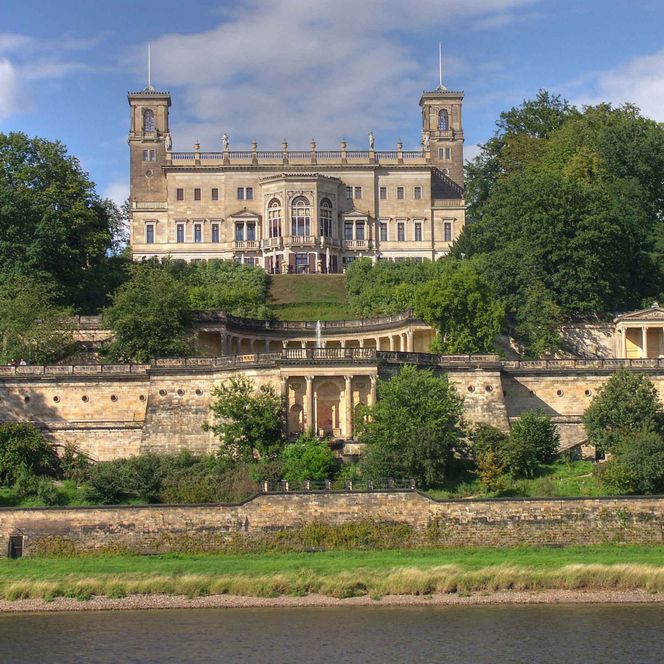 Historisches Schloss mit Türmen, umgeben von Bäumen und einem Fluss im Vordergrund.