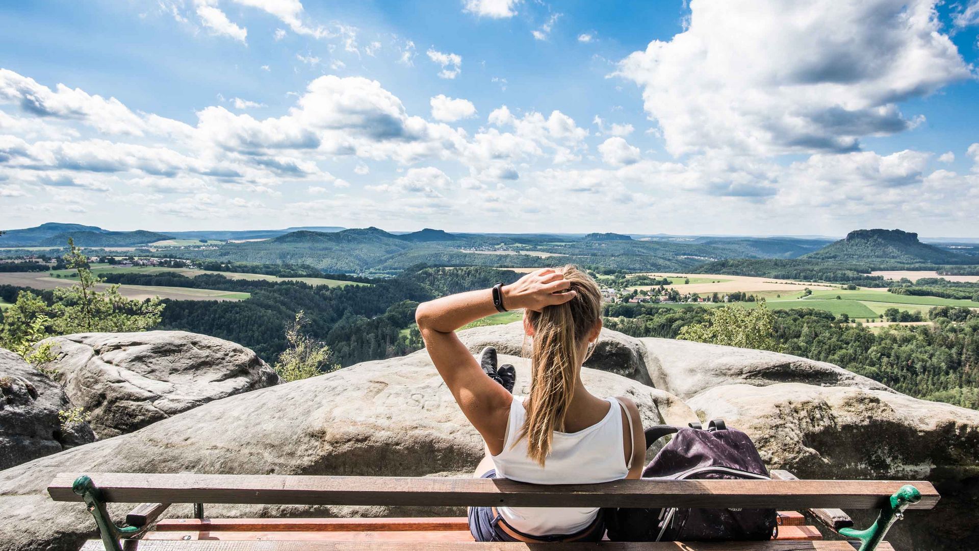 Frau mit langen Haaren sitzt auf einer Bank und blickt auf eine weite Landschaft mit Hügeln und Wolken.