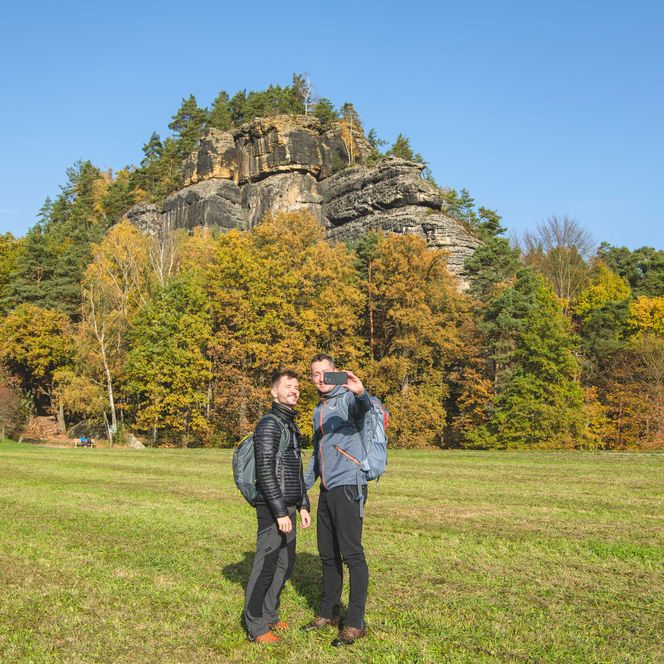 Zwei Personen stehen auf einer Wiese vor einem bewaldeten Hügel mit herbstlichen Bäumen und dem Fels Rauenstein im Hintergrund.