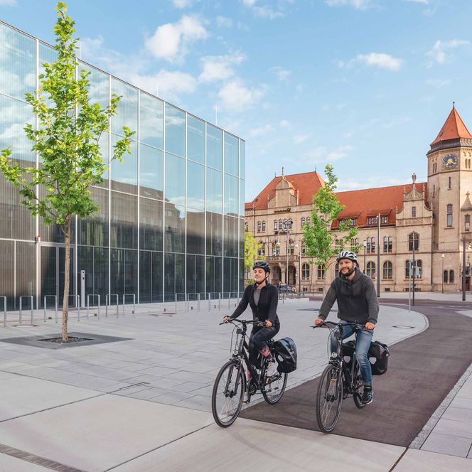 Zwei Kinderwagen stehen auf einem gepflasterten Weg, im Hintergrund ein historisches Gebäude und Bäume.