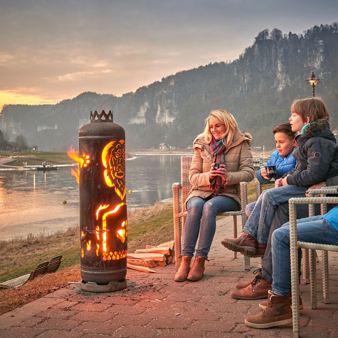 Familie sitzt um einen Feuerkorb am Ufer der Elbe in Rathen bei Sonnenuntergang.