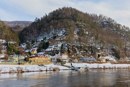 Schneebedeckte Hügel mit Bäumen, darunter ein Dorf am Wasser mit Booten.