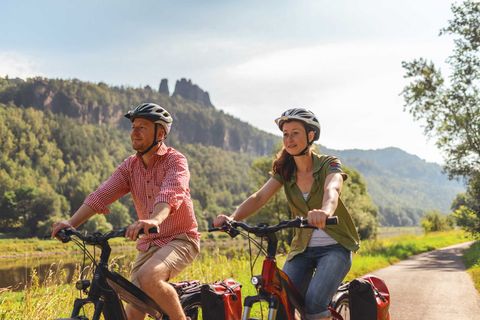Zwei Radfahrer auf dem Elberadweg in der Sächsischen Schweiz, im Hintergrund die bewaldeten Elb-Hänge und Felsformationen des Elbsandsteingebirges.