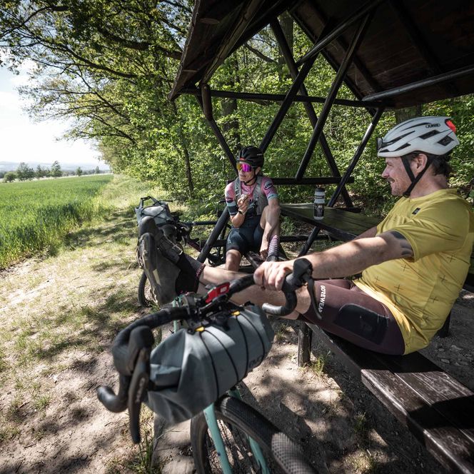 Zwei Radfahrer sitzen unter einem Pavillon, umgeben von Bäumen und Wiesen.
