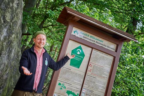 Mann in Jacke zeigt auf einen Informationsstand mit Wanderkarten und Hinweisen im Wald.