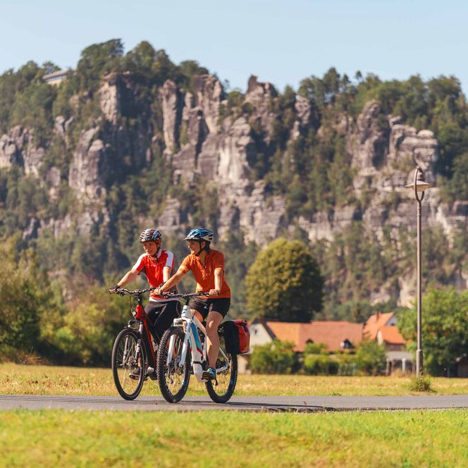 Zwei Radfahrer in roten Trikots fahren auf einem Weg, im Hintergrund sind Felsen und Bäume zu sehen.