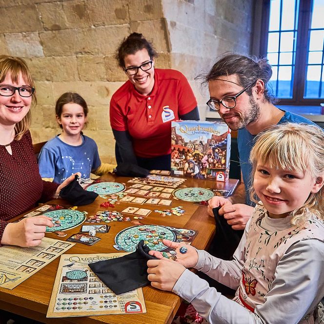 Familie spielt gemeinsam an einem Tisch mit einem Brettspiel in einem gemütlichen Raum.