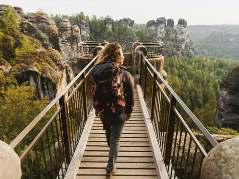 Frau mit Wanderrucksack von hinten läuft auf Holzbrücke über Felsenschlucht, im Hintergrund Wälder.